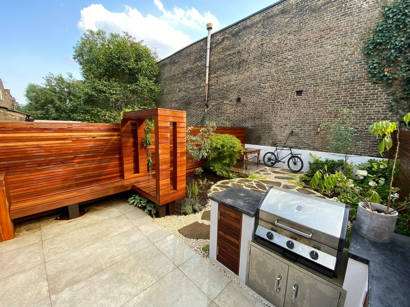 A courtyard with a cedar slatted bench and screen, stone paving, planting, and a built-in stainless steel barbecue.