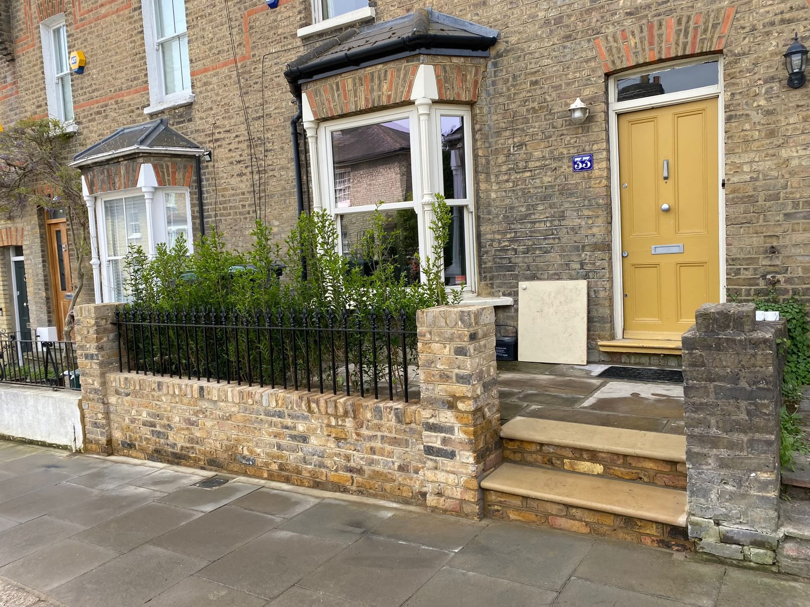 A brick terrace front garden with a low brick wall, black railings, dense shrubs, and a yellow front door.