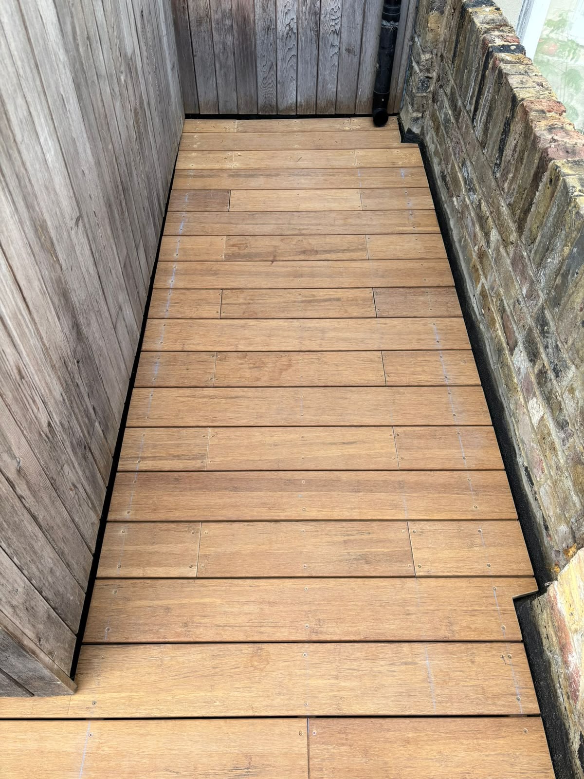 A narrow side return laid with warm timber decking between a fence and a brick wall.