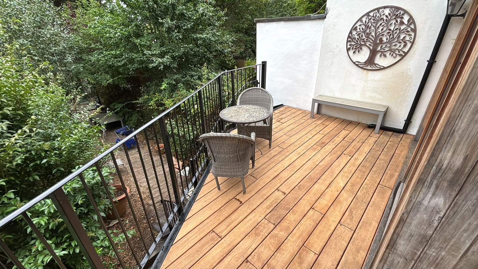 An upper deck in warm boards, with wicker chairs, a small table, and a metal tree-of-life panel on a white wall.