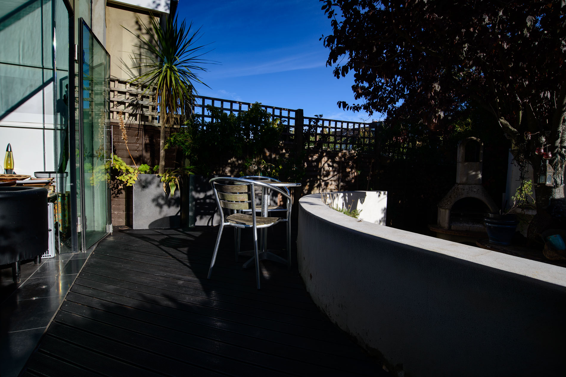 A dark timber deck beside a curved white wall, with metal chairs, glass doors, and a shaded garden beyond.